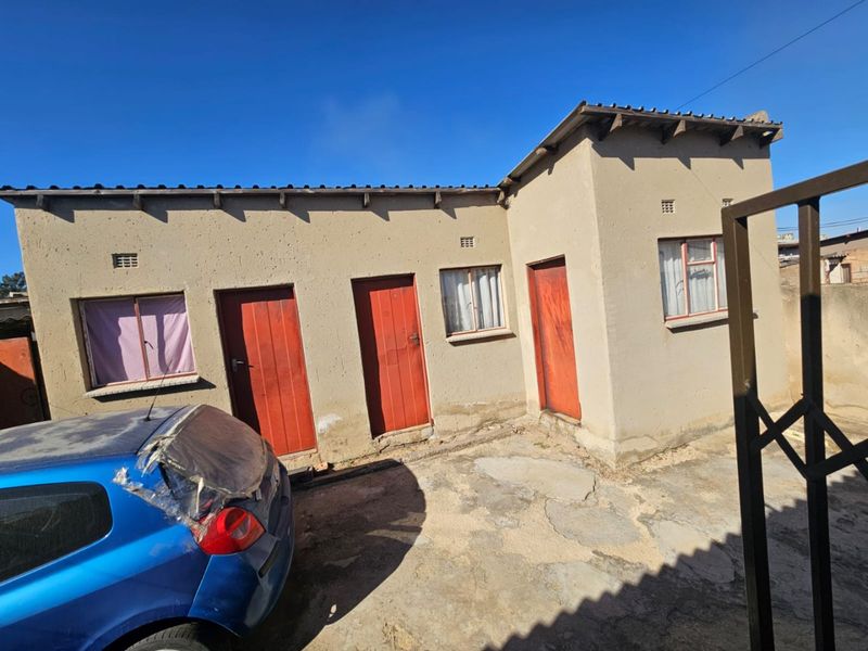 Rear exterior view of a single-story residential building featuring plastered walls, multiple timber access doors, and concrete paved yard area.