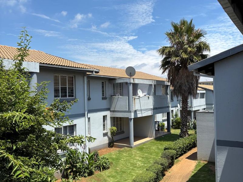 View of the apartment complex rear elevation featuring landscaped gardens, paved walkways, and private balconies under a clear blue sky.