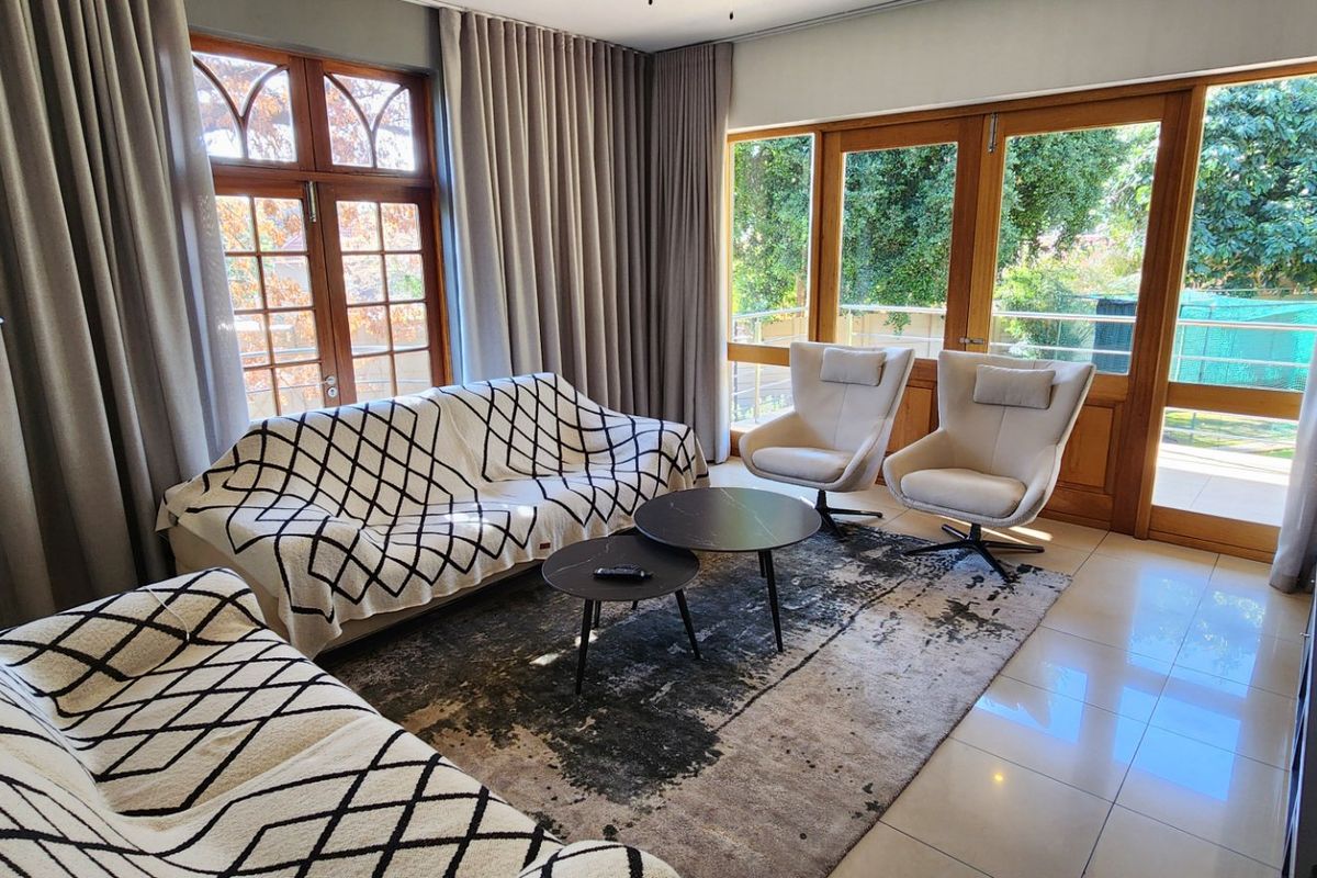 A bright living room featuring large timber-framed glass doors and windows, tiled flooring, and recessed ceiling lighting, offering a pleasant view of the garden.