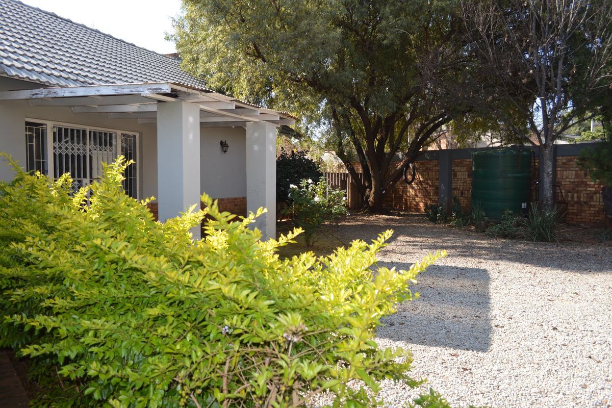 Rear exterior view of the house featuring a covered patio, gravel yard, mature trees, and a brick boundary wall with a water storage tank.