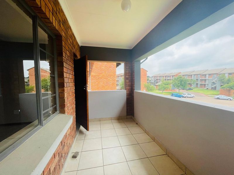 A covered balcony featuring tiled flooring and a brick exterior wall, offering an open view of the surrounding apartment complex.