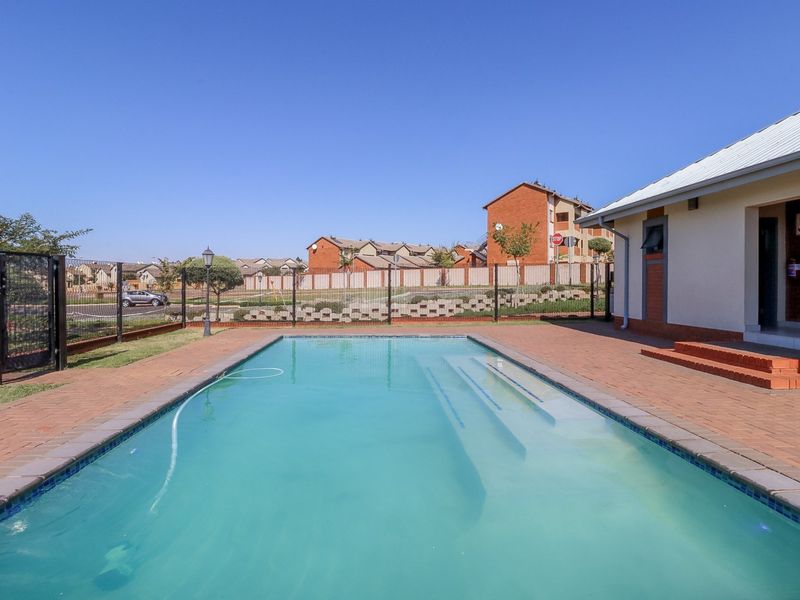 A well-maintained outdoor swimming pool area featuring brick paving, perimeter fencing, and clear views of the surrounding residential complex.