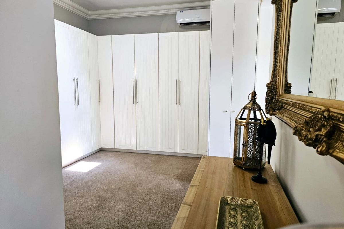 A well-maintained bedroom featuring extensive white built-in wardrobes, neutral carpeting, and a wall-mounted split-system air conditioning unit.