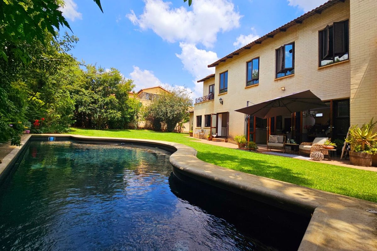 A well-maintained rear exterior view featuring a curved swimming pool, manicured lawn, and a paved patio area adjacent to the main house structure.