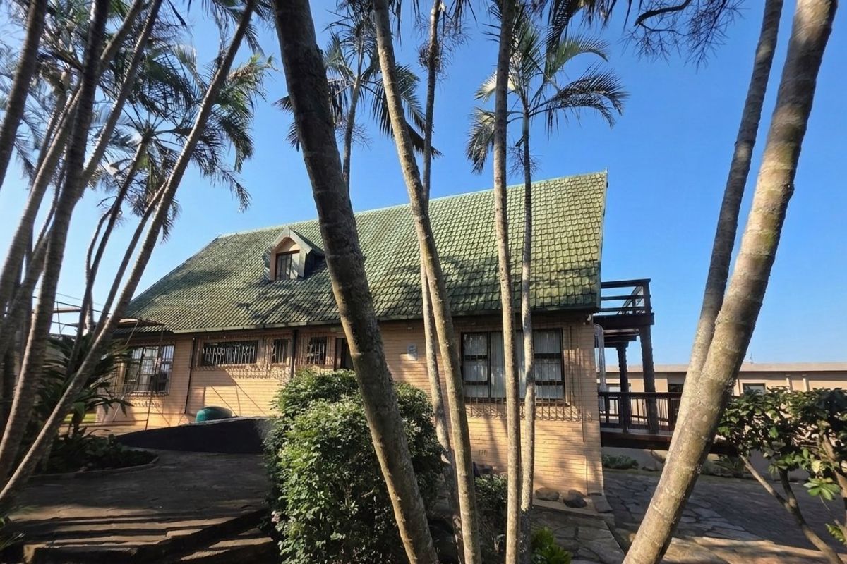 Rear view of a brick house featuring a pitched tiled roof, a raised wooden deck, and a paved patio area surrounded by mature trees.