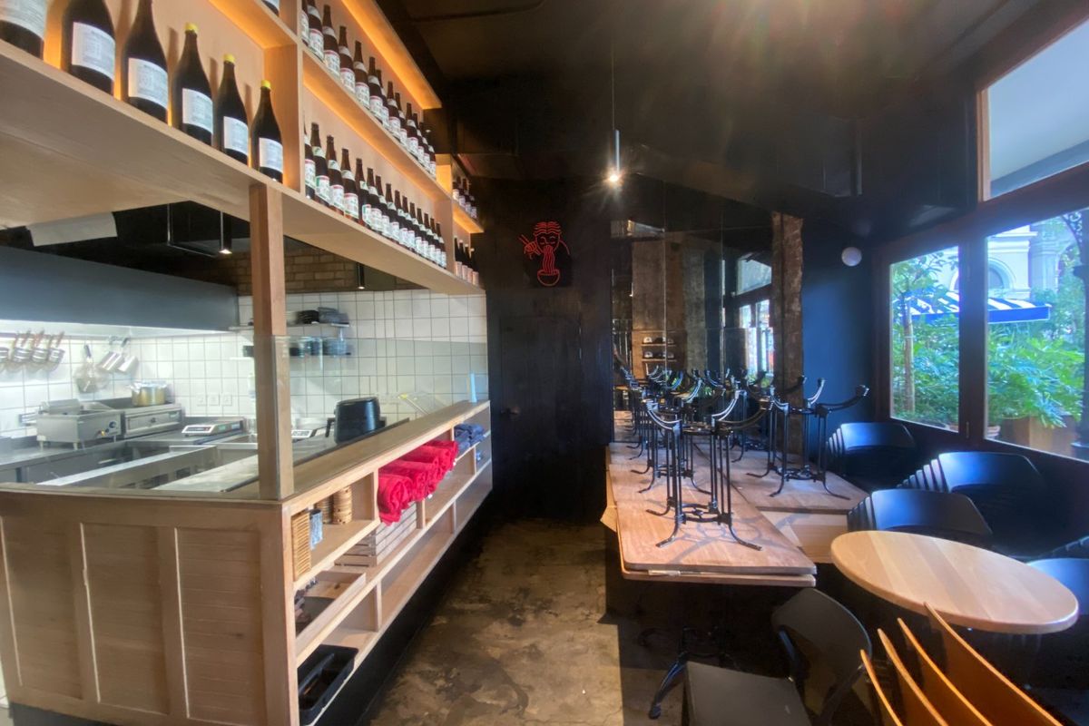 A modern commercial restaurant space featuring an open-plan layout with a built-in kitchen service area, concrete flooring, and large windows overlooking the exterior.