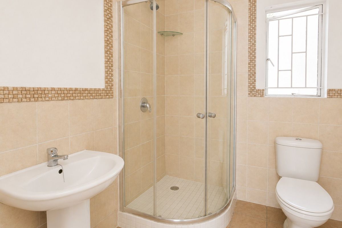 A functional bathroom featuring a corner shower enclosure, pedestal basin, and toilet with neutral wall tiling and decorative mosaic accents.