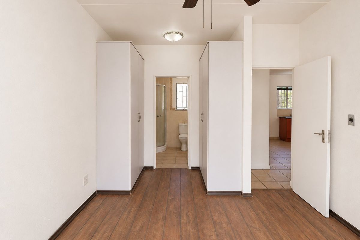 A bedroom featuring built-in cupboards, wood-look laminate flooring, and a ceiling fan, with an adjoining en-suite bathroom and access to a tiled hallway.