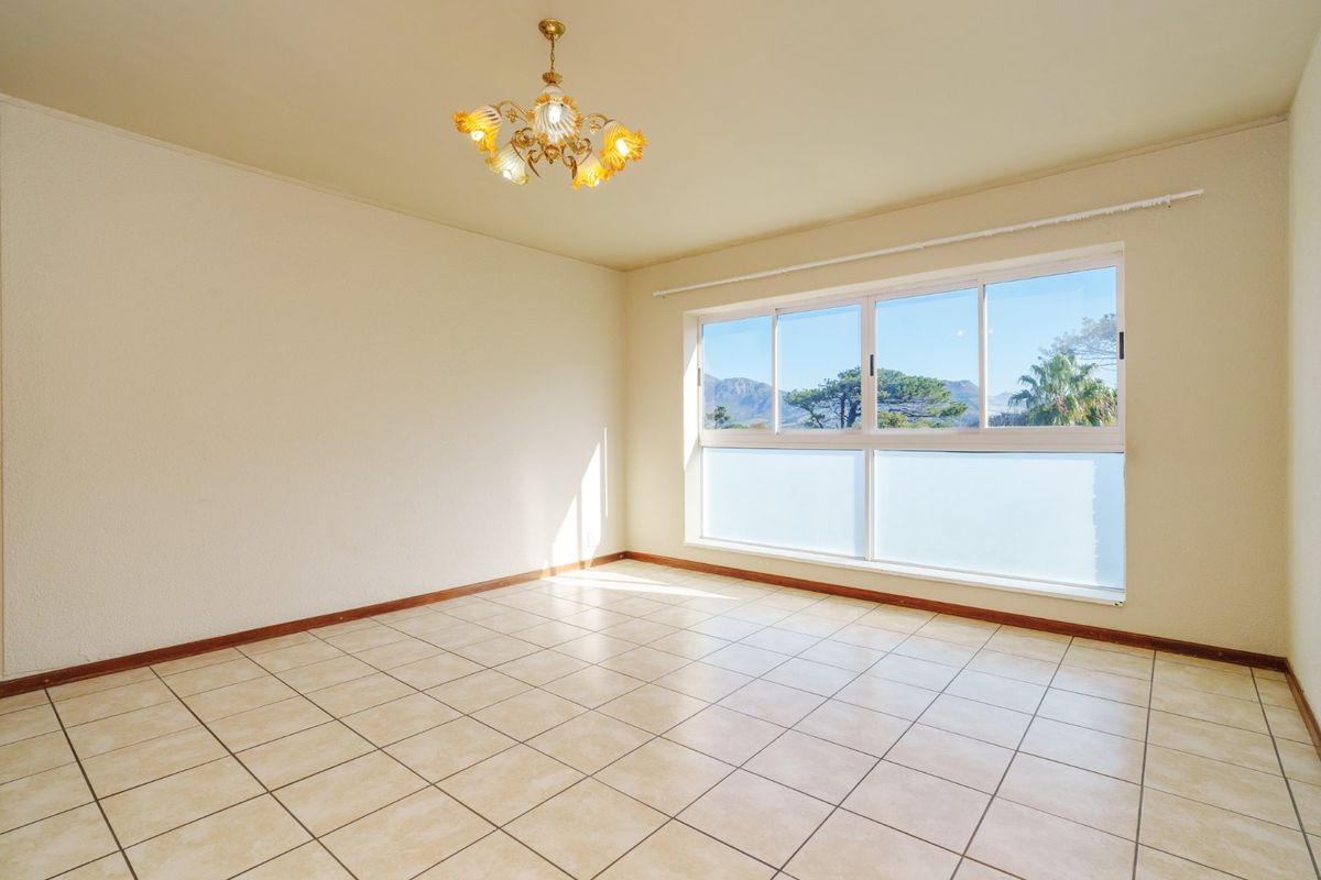 A bright, empty living room featuring neutral tiled flooring, a large window with mountain views, and a classic chandelier fixture.