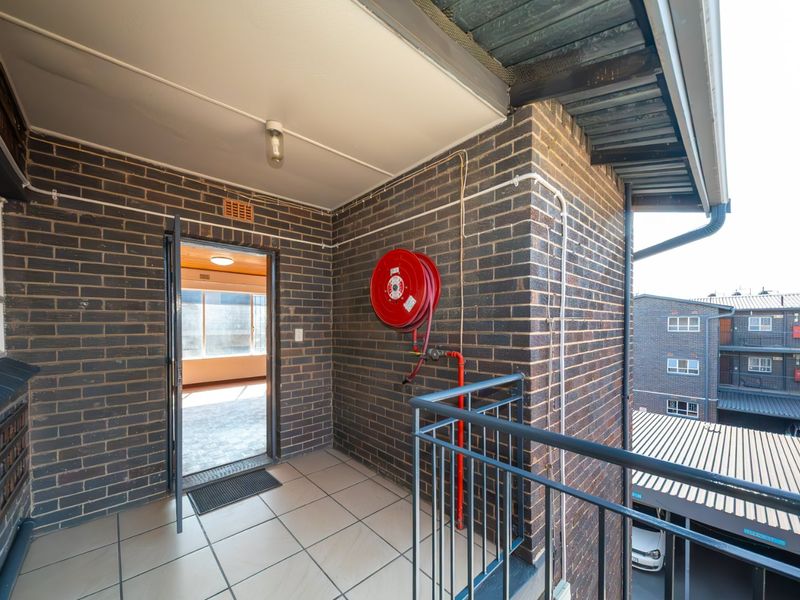 A functional apartment balcony featuring exposed brick walls, tiled flooring, and a wall-mounted fire hose reel, providing access to the interior living space.