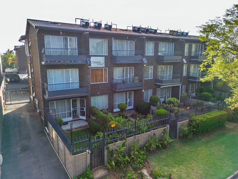 Exterior view of Desnor Court apartment complex featuring a multi-story brick facade with balconies, solar water heating systems, and secure perimeter fencing.
