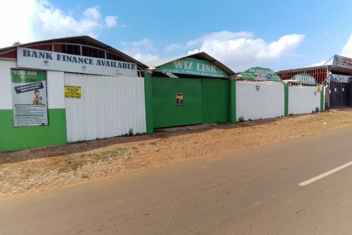 Commercial vehicle dealership frontage featuring corrugated metal fencing, secure gated access, and prominent business signage along a paved street.