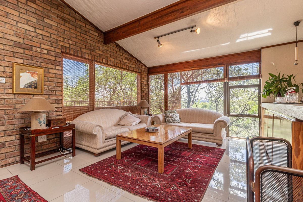 A sunlit living room featuring exposed brick walls, timber ceiling beams, large windows with garden views, and polished tiled flooring.