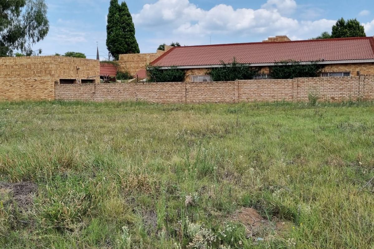 A view of a vacant residential plot featuring a grassy area with a brick boundary wall and adjacent residential building in the background.
