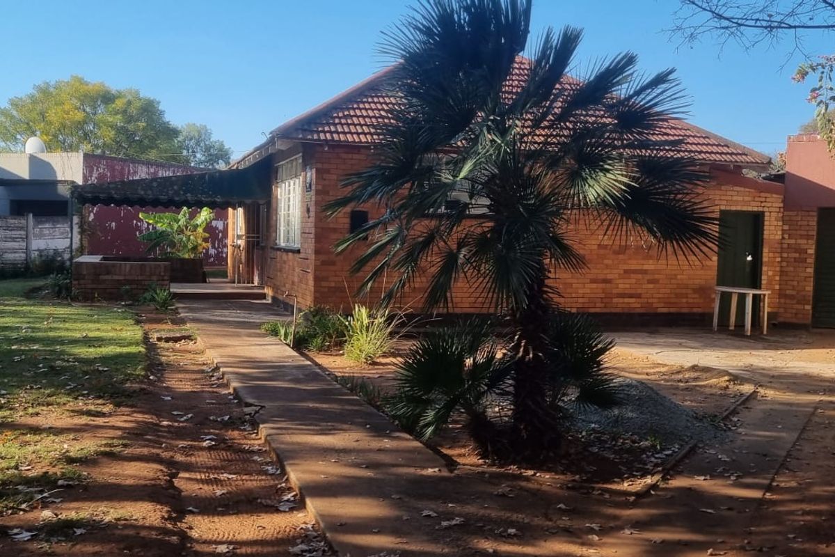 Rear view of a brick house featuring a tiled roof, concrete walkway, and attached garage, set within a garden area with a boundary wall.