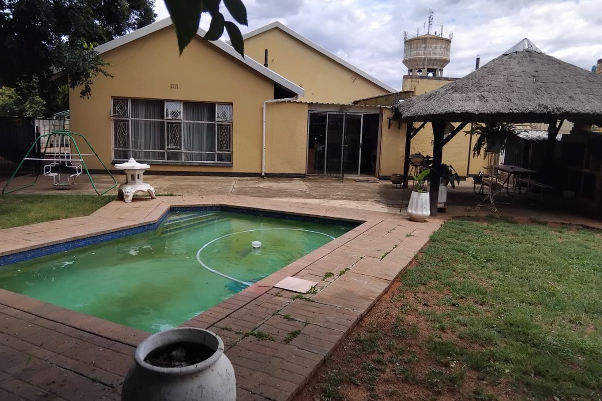 Rear exterior view featuring a swimming pool, paved patio area, and a traditional thatched lapa structure in the garden.
