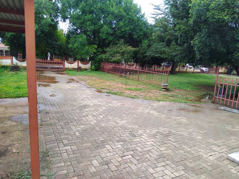 A paved driveway leading to a gated entrance, featuring metal perimeter fencing and mature trees in the surrounding garden area.