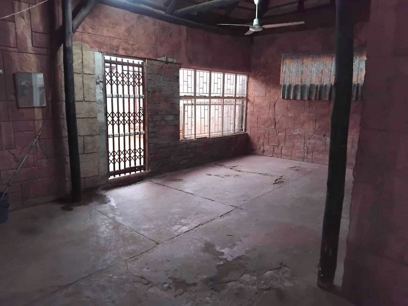 An empty living area featuring exposed timber beams, stone-effect wall cladding, and a security gate, requiring significant cosmetic updates to the flooring and walls.