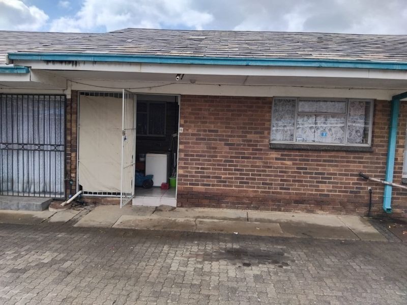 Front exterior view of a brick apartment unit featuring a security gate entrance, paved driveway, and tiled roof structure.