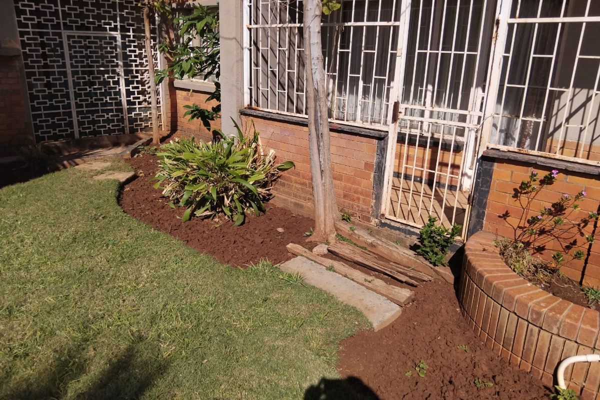 A rear garden area featuring a brick facade, security gates, and a built-in brick planter box with established lawn and garden beds.
