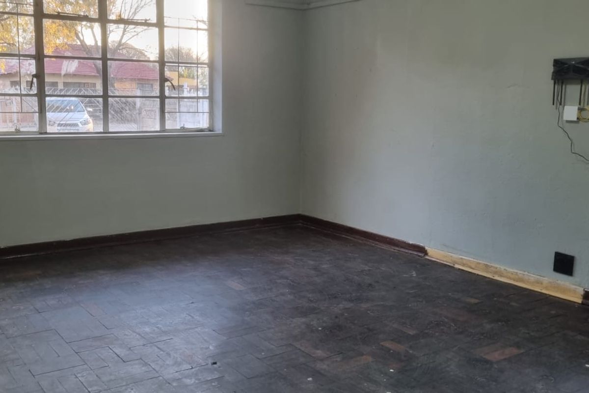 An empty living room featuring original parquet flooring, steel-framed windows with security bars, and neutral wall finishes in need of cosmetic attention.