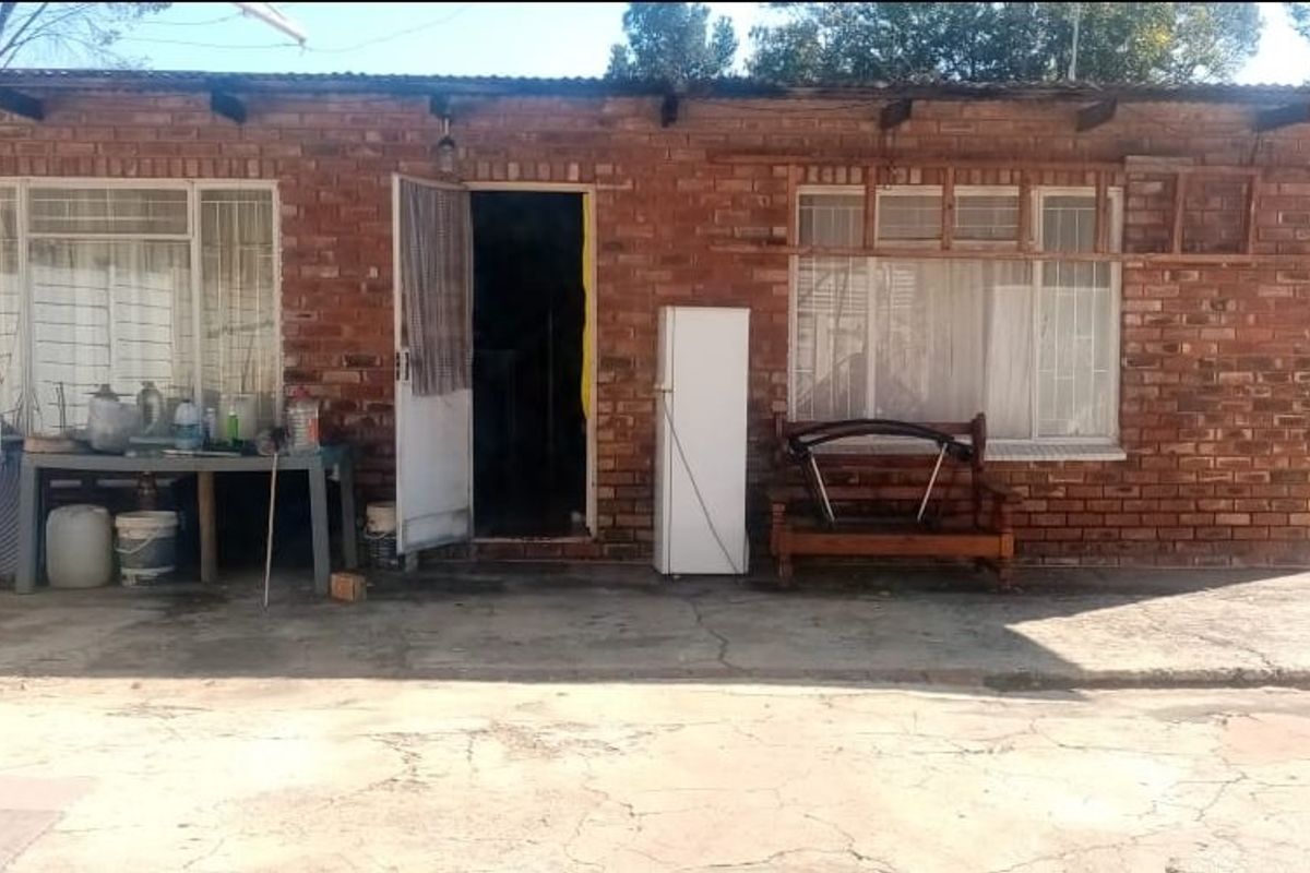 Rear exterior view of a single-story brick house featuring a concrete patio area, security window bars, and a corrugated metal roof.