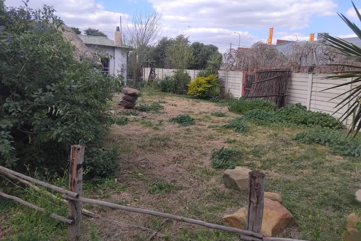 A rear garden area featuring a precast concrete boundary wall, a thatched roof structure, and established garden beds requiring maintenance.
