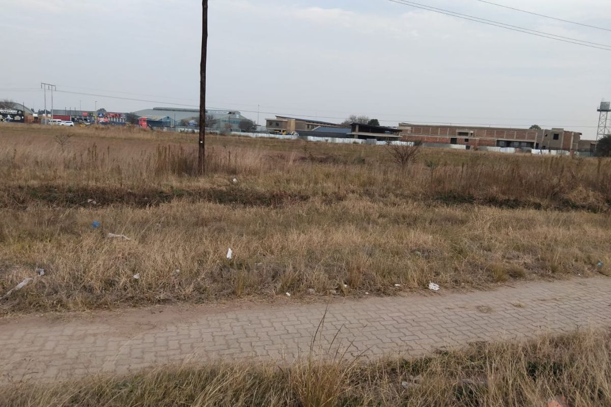 A view of a vacant residential plot featuring a paved walkway and utility pole, with surrounding neighborhood development visible in the background.