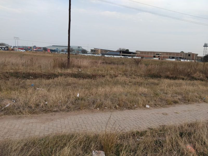 A view of a vacant residential plot featuring a paved walkway and utility pole, with surrounding neighborhood development visible in the background.