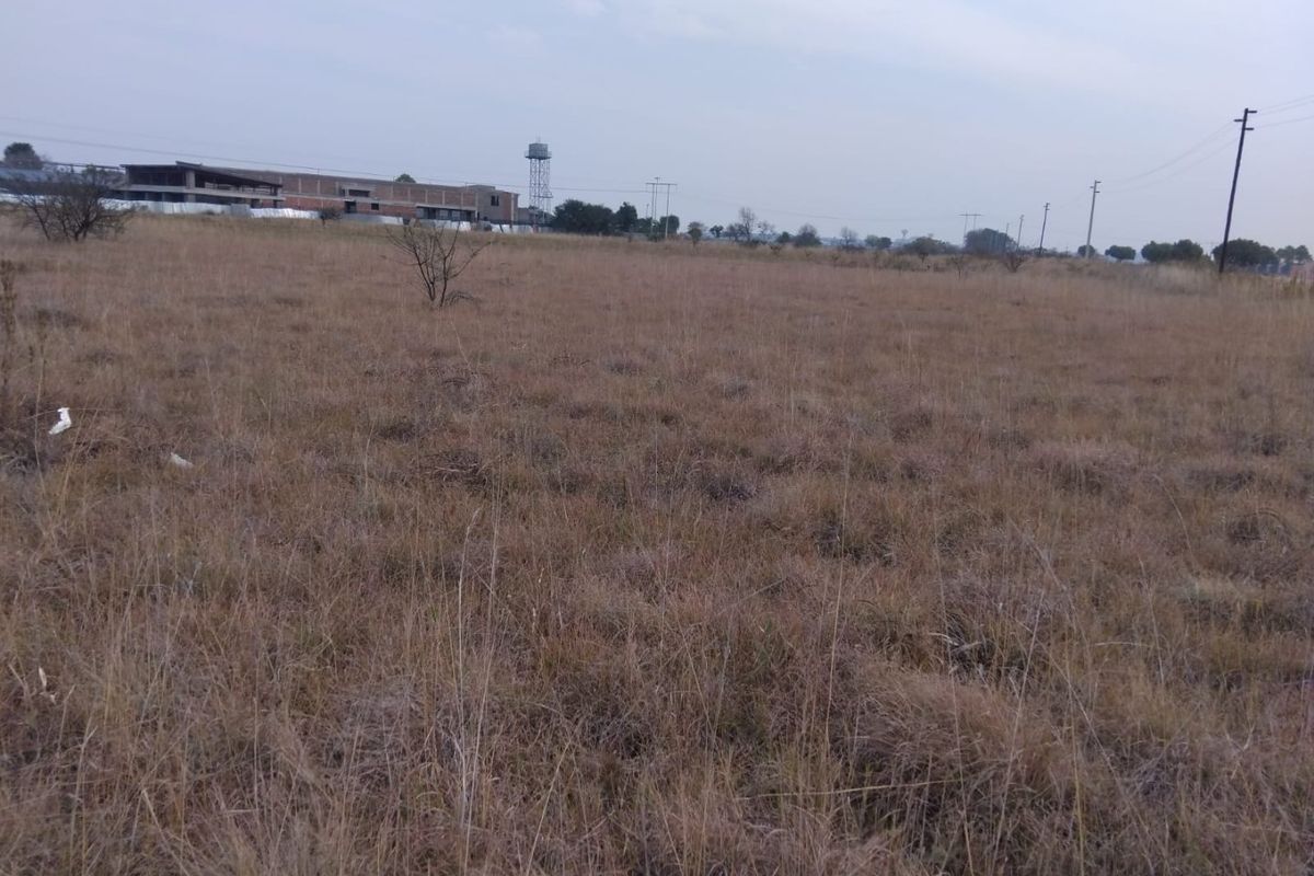 A wide, open vacant residential plot featuring dry grass, with distant views of a water tower and utility infrastructure.