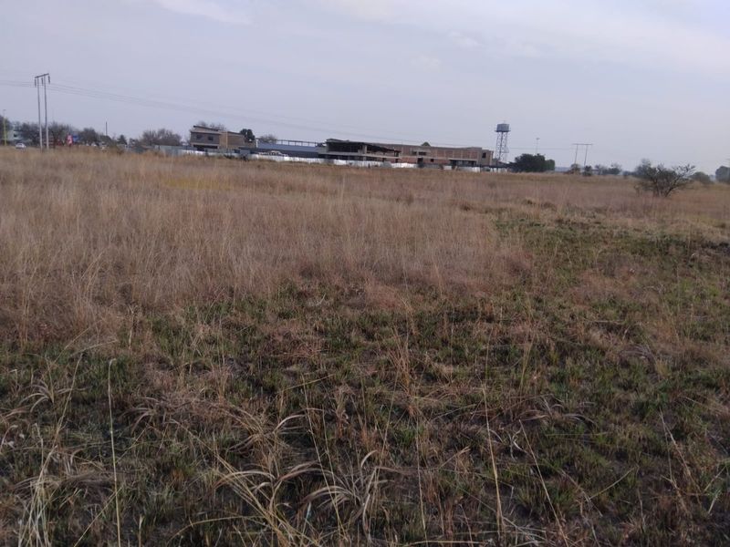 A view of a vacant residential plot featuring dry grass and surrounding infrastructure in the distance.