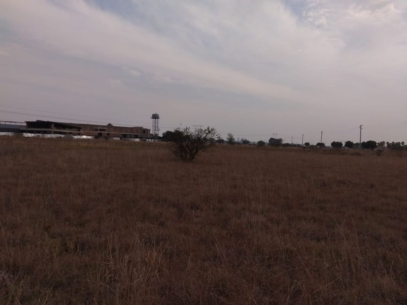 A wide view of a vacant residential plot featuring dry grass, distant power lines, and a water tower in the background.