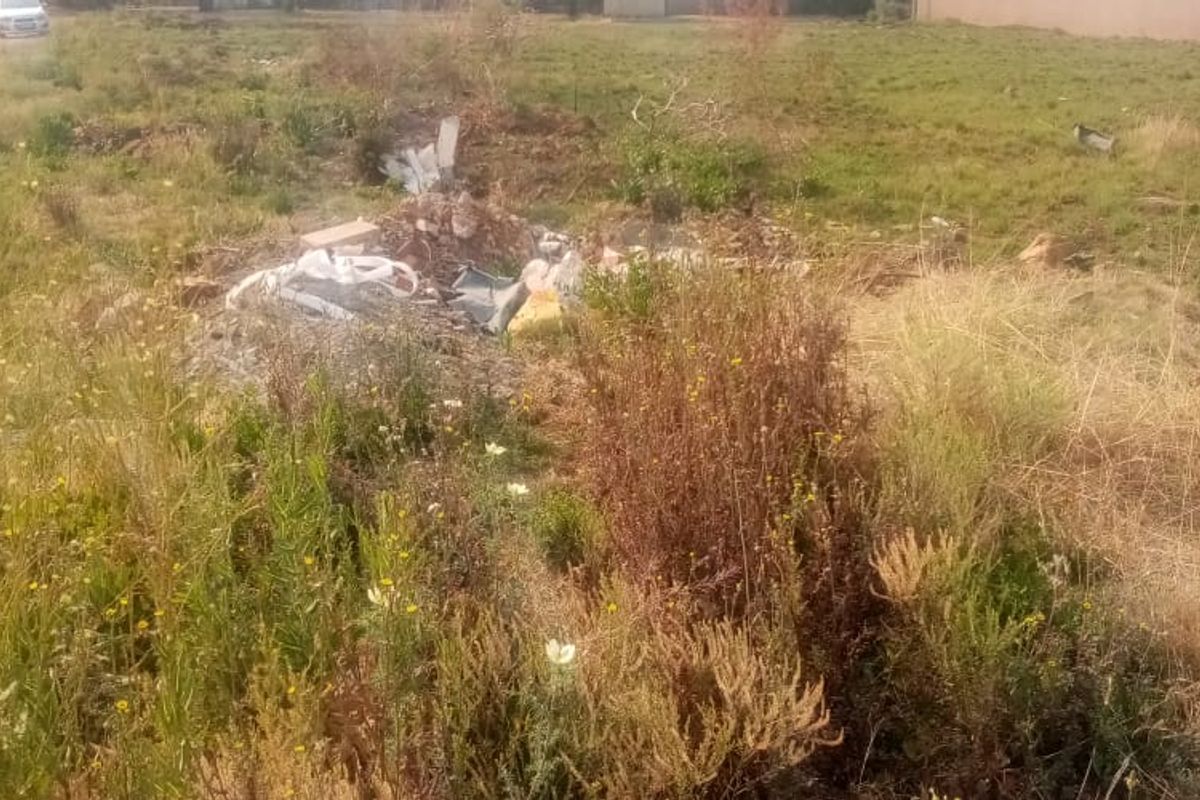 A view of a vacant residential plot featuring overgrown grass and accumulated debris on the site.