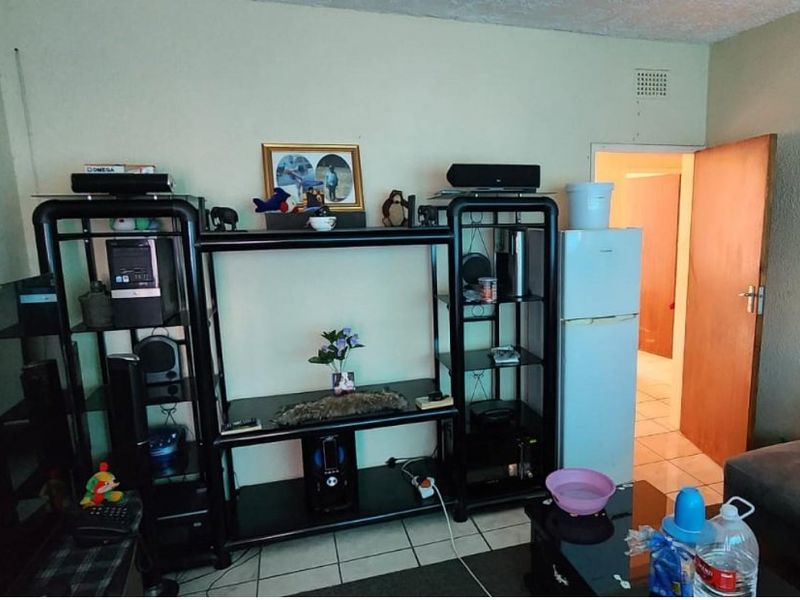 A living room area featuring tiled flooring and a ventilation grille, currently occupied by personal storage units and furniture.