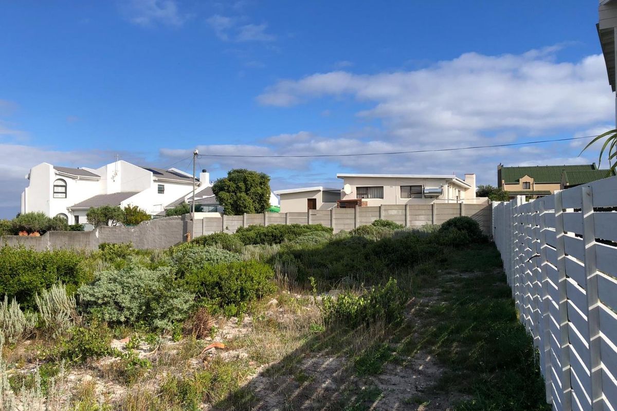 A vacant residential plot featuring natural coastal vegetation, enclosed by a precast concrete boundary wall and a white timber slat fence.
