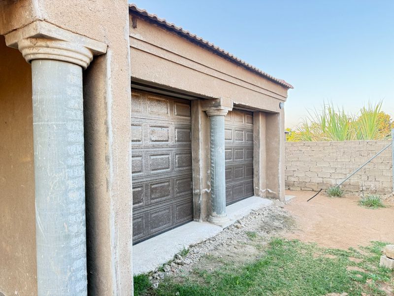 A double garage exterior featuring decorative columns and brown sectional doors, currently showing unfinished plasterwork and surrounding ground works.