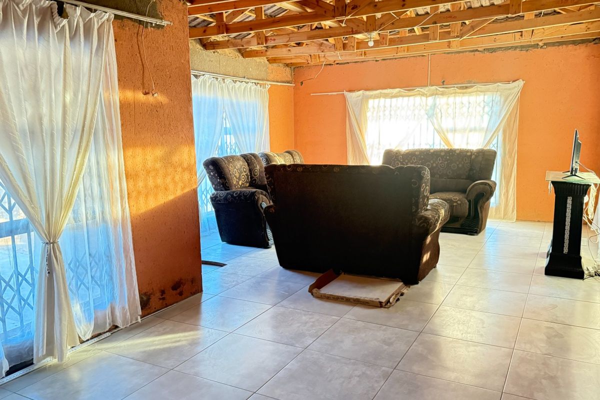 Spacious living area featuring exposed timber roof trusses, ceramic tiled flooring, and security gates, requiring cosmetic finishing to walls and ceilings.
