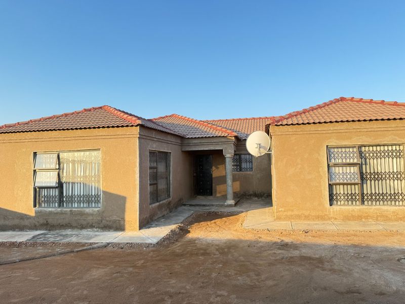Front elevation of a single-story house featuring a tiled roof, security gates, and a concrete entrance column, with unfinished surrounding ground.