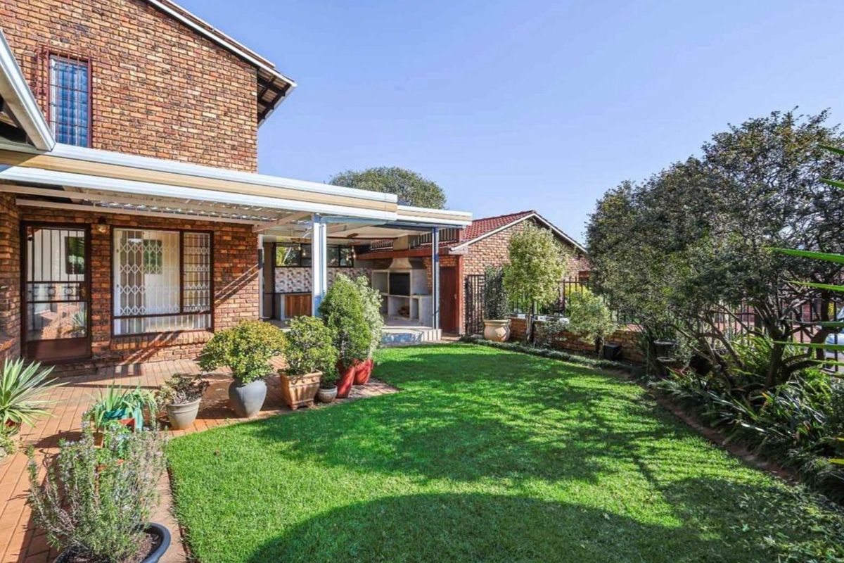 Rear view of the townhouse featuring a brick facade, a covered patio with a built-in braai, and a well-maintained garden with a lawn.