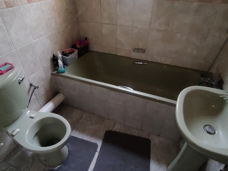 A dated bathroom featuring a green bathtub, matching pedestal basin, and toilet, with full-height wall tiling and an exposed plumbing pipe.