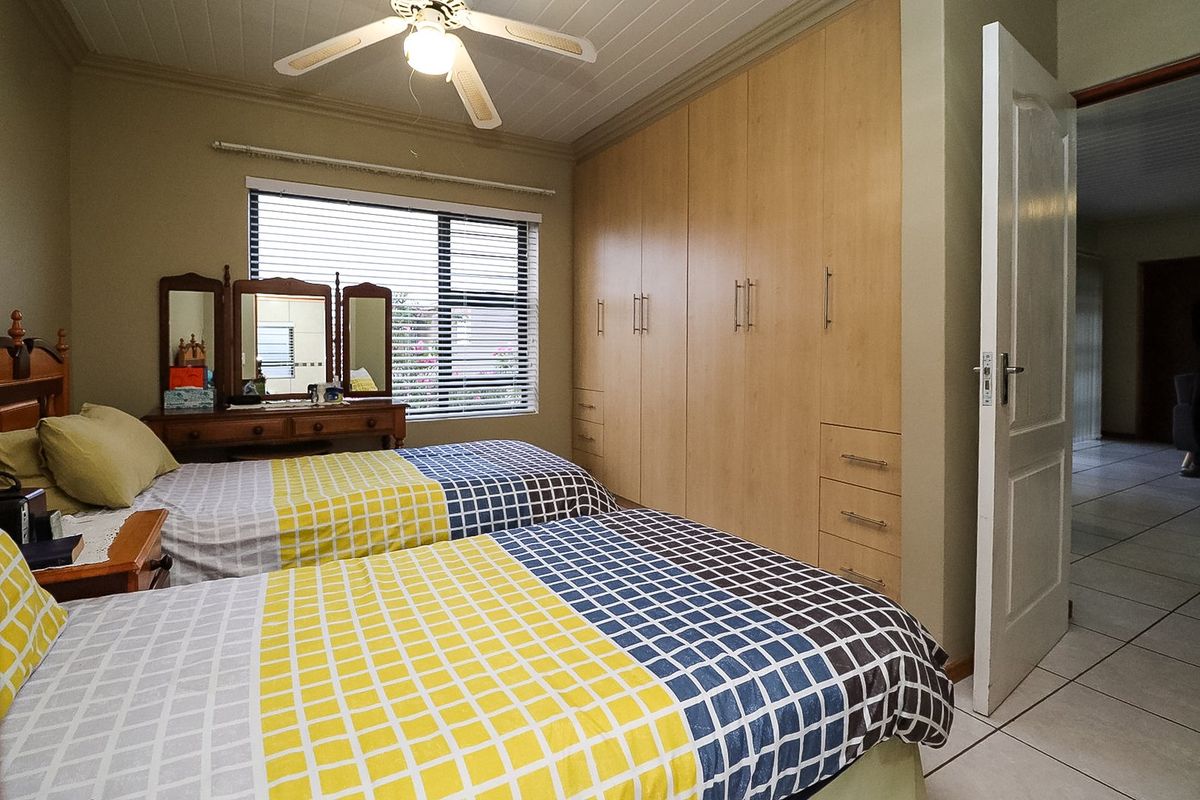 A well-maintained bedroom featuring built-in wooden cupboards, tiled flooring, a ceiling fan, and large windows with venetian blinds.
