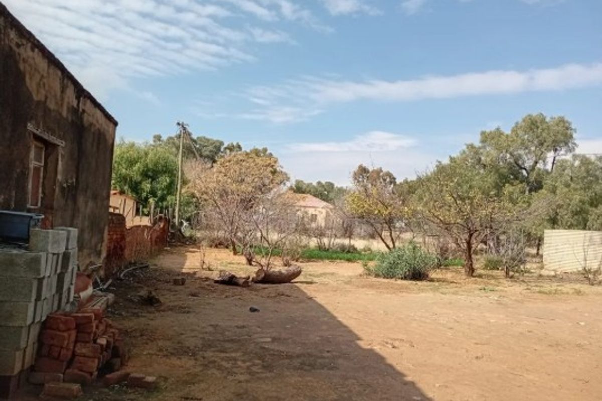 A view of the rear yard area featuring an existing structure wall, open ground, and mature trees on the residential plot.