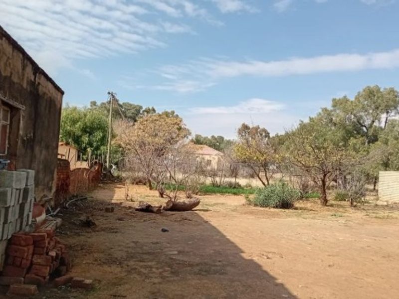 A view of the rear yard area featuring an existing structure wall, open ground, and mature trees on the residential plot.