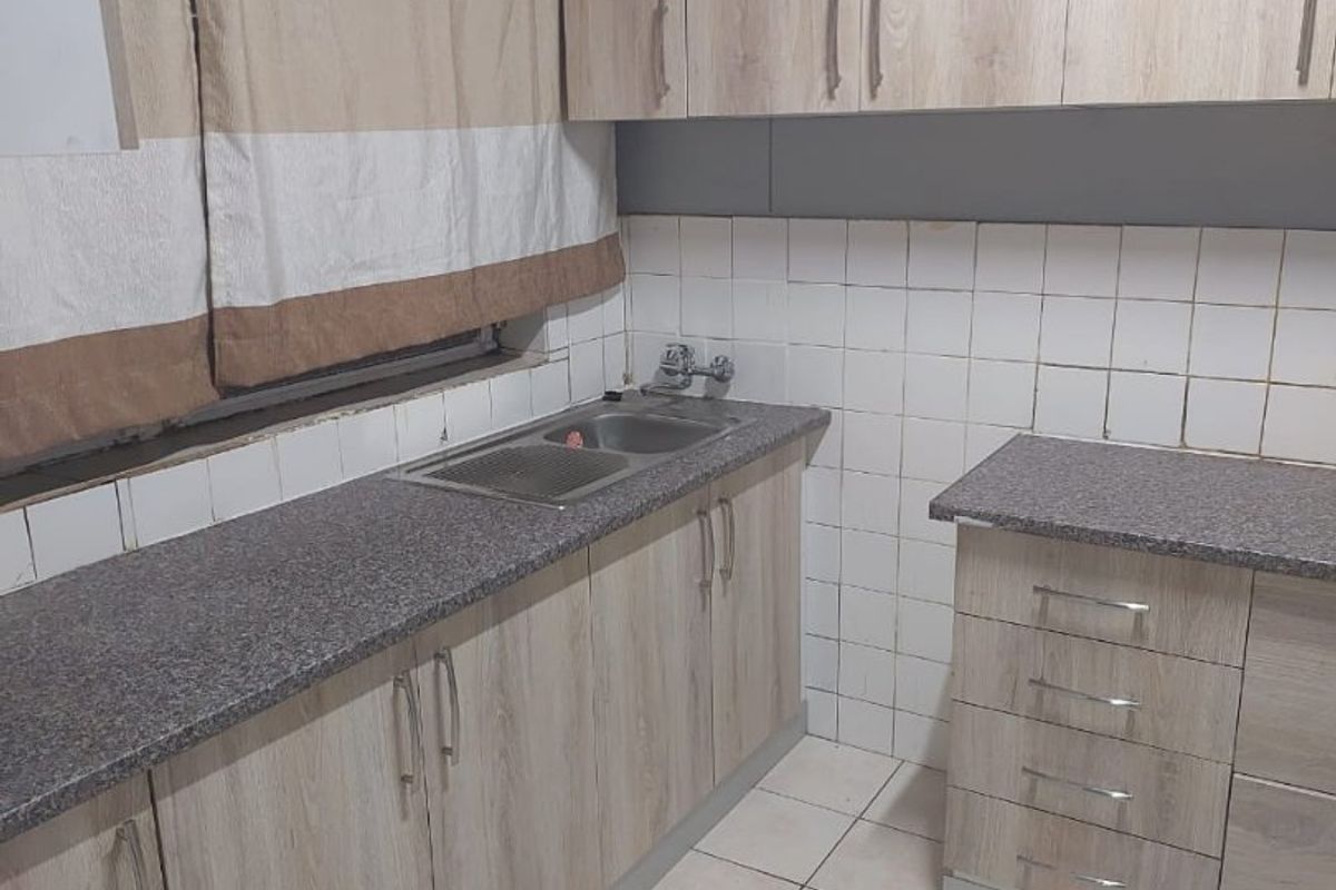 A functional kitchen featuring light wood-look cabinetry, granite-effect countertops, a stainless steel sink, and tiled splashbacks with neutral floor tiling.