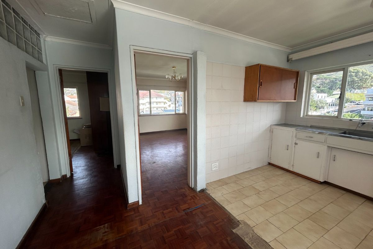 An empty kitchen area featuring dated cabinetry, tiled splashbacks, and a mix of parquet and ceramic tile flooring, leading into an adjacent living space.