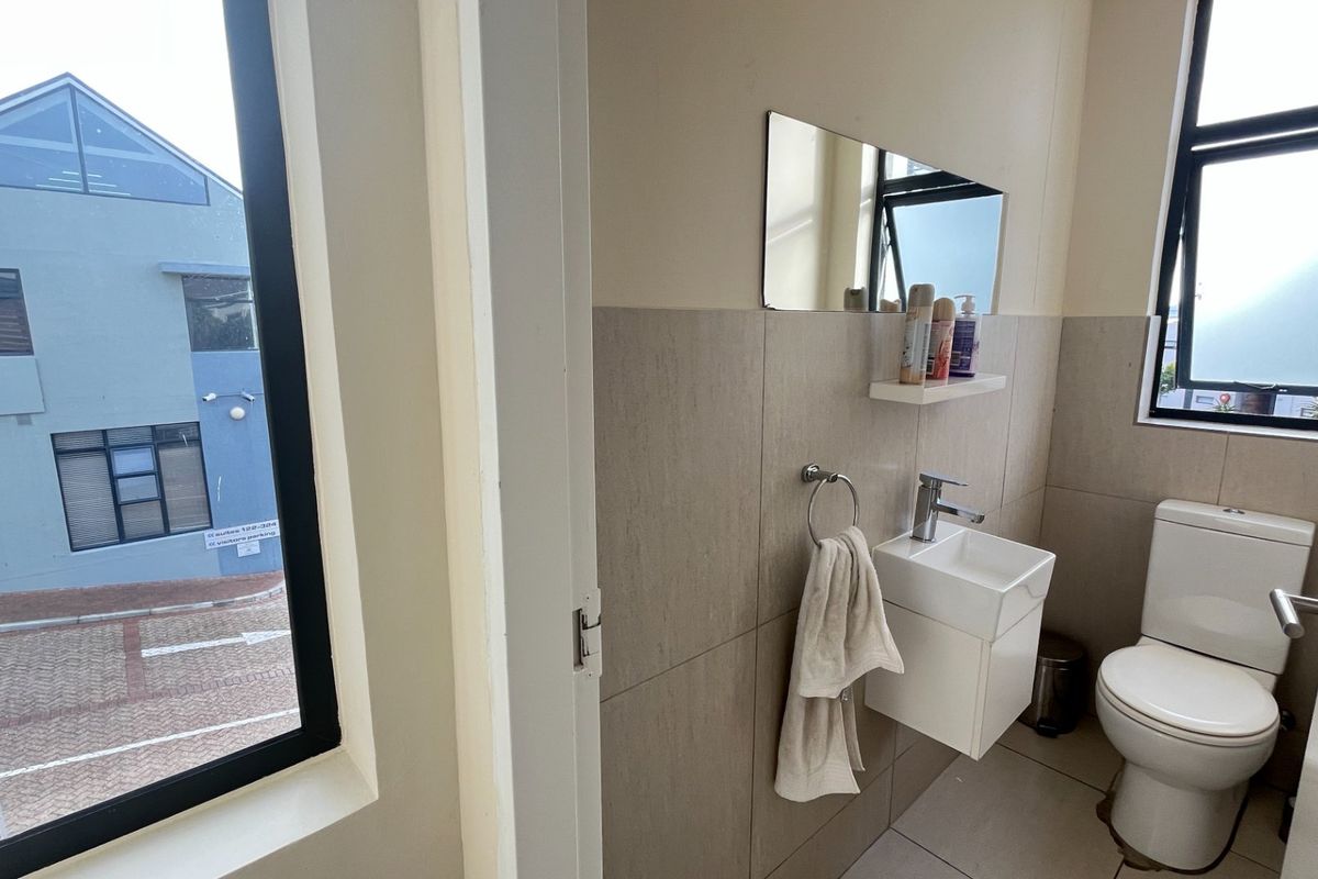 A compact commercial bathroom featuring a wall-mounted basin, toilet, and neutral tiled finishes with natural light from a side window.