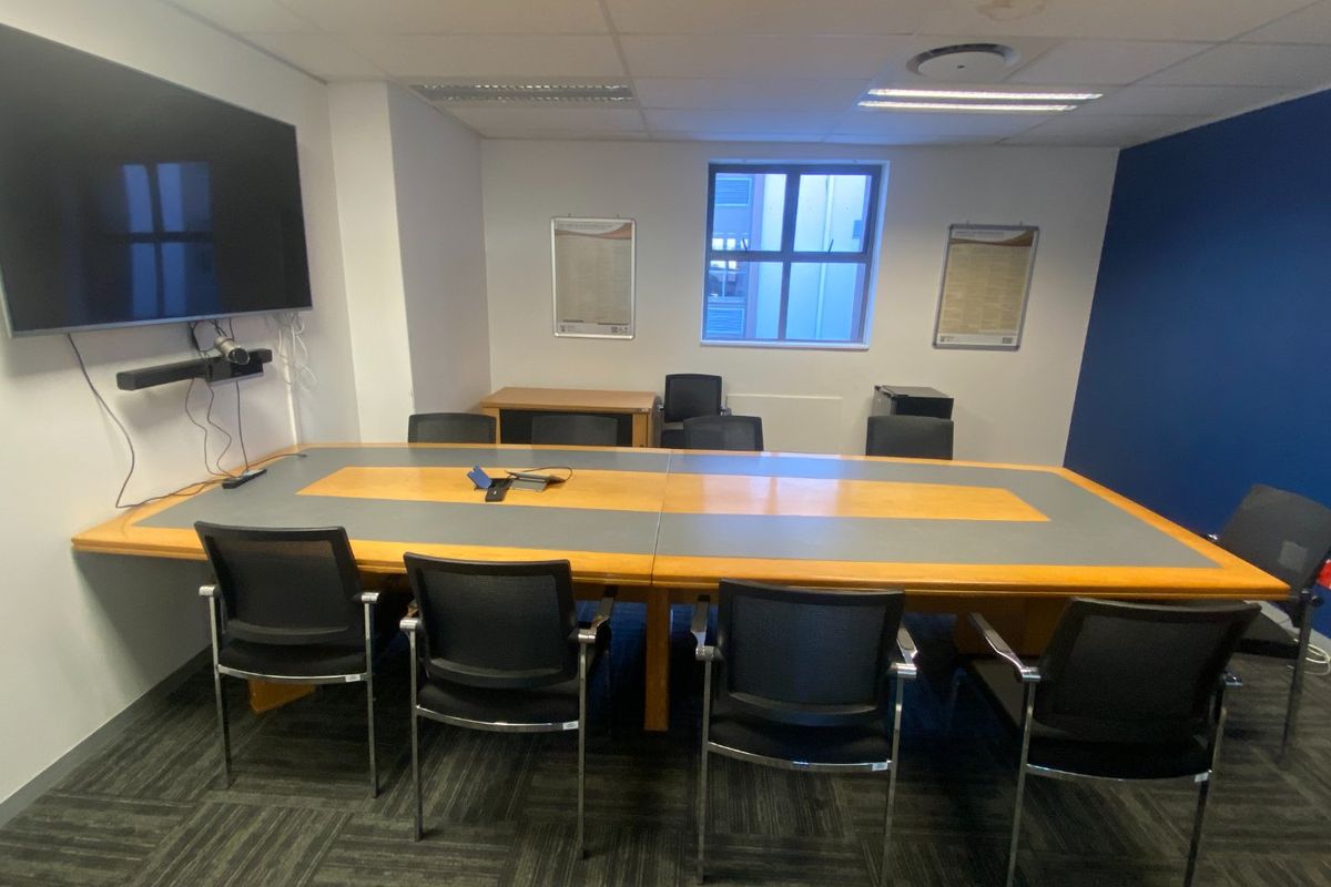 A functional office meeting room featuring a large conference table, carpeted flooring, suspended ceiling, and a wall-mounted display screen.