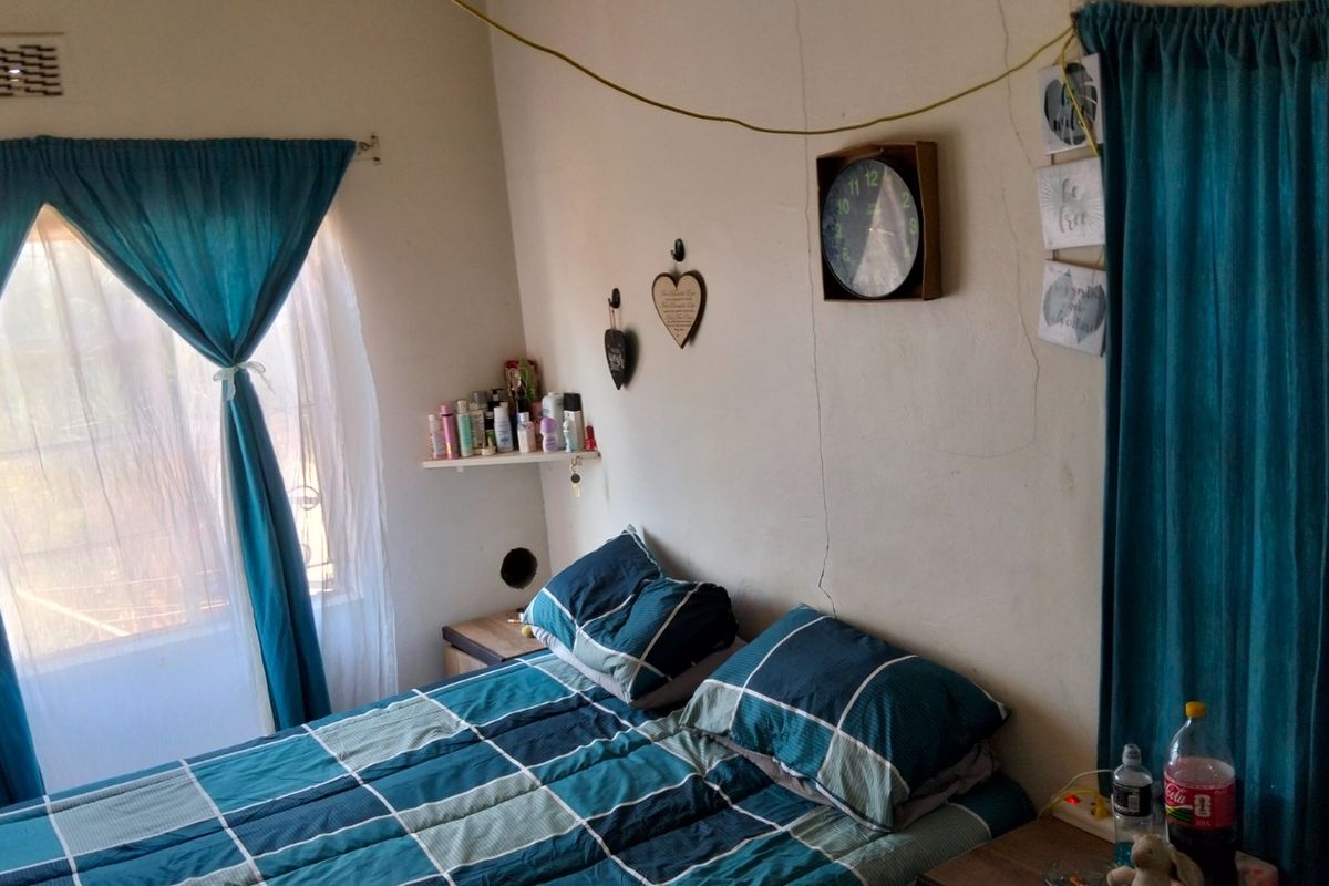 A bedroom featuring a window with curtains and visible wall damage, including significant cracks and a hole in the wall surface.