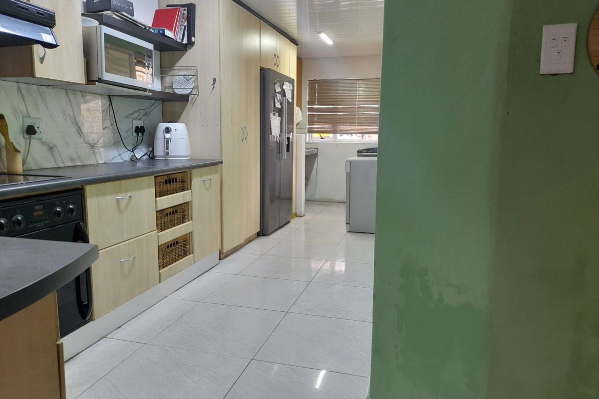 A functional kitchen featuring light wood-finish cabinetry, tiled flooring, and built-in appliances, with an adjacent utility area visible in the background.