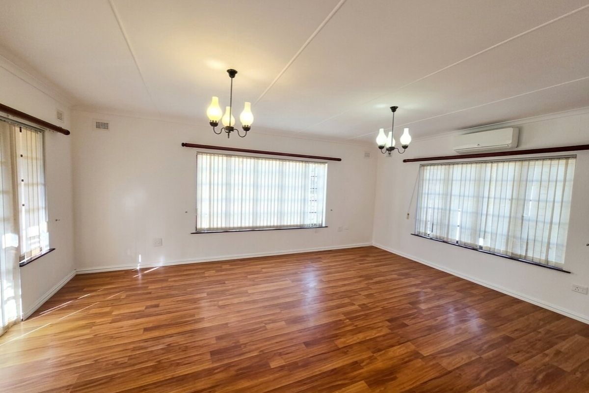 Spacious, light-filled living room featuring warm-toned laminate flooring, vertical blinds, and a wall-mounted split-system air conditioner.